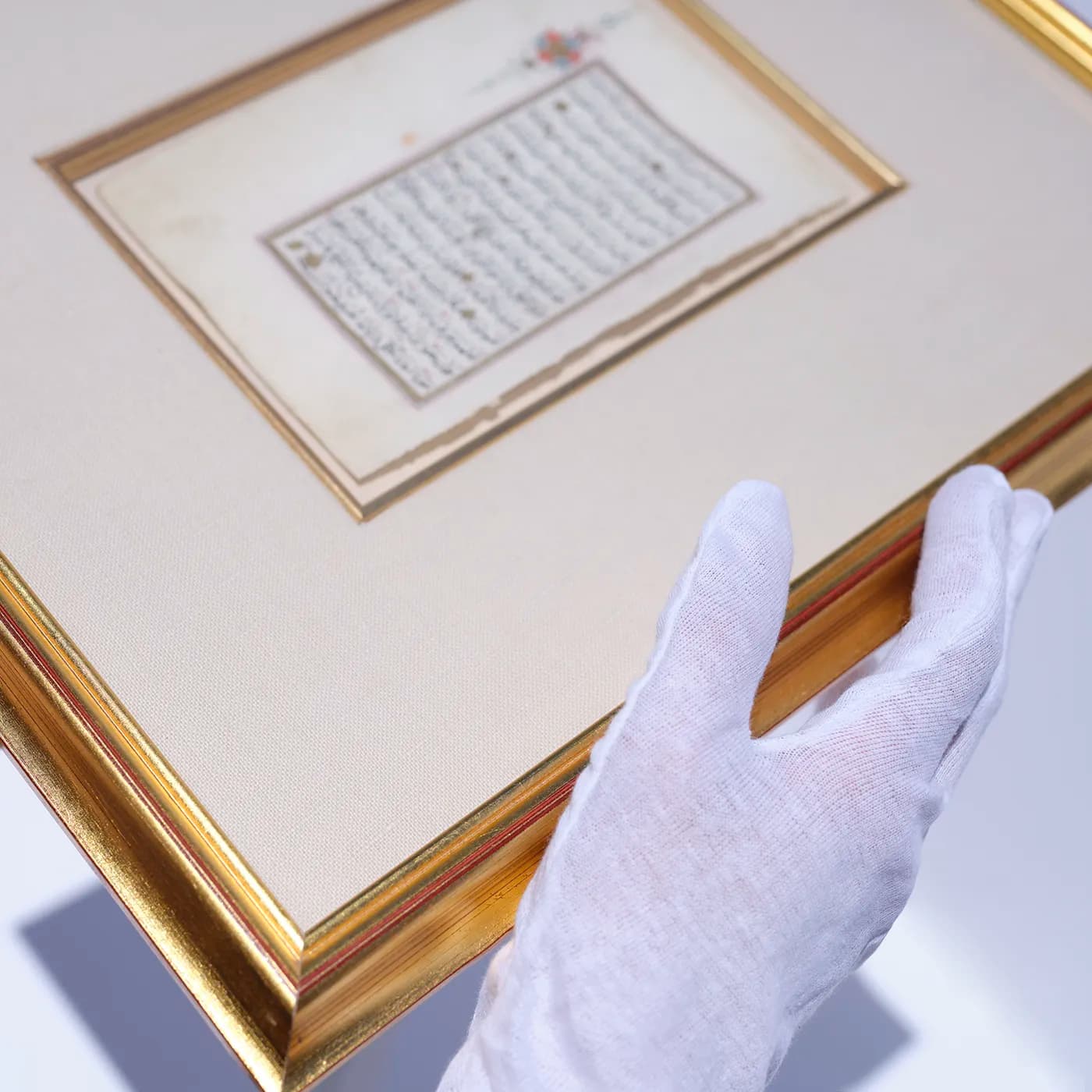 White-gloved conservator examining a framed antique manuscript with gold-leaf border